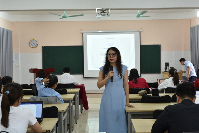 A person in a blue dress holding a microphone in front of a classroom

Description automatically generated